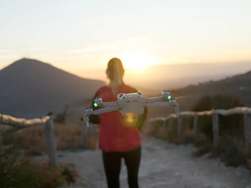 Eine Pilotin hält die aufgeklappte DJI Mini 4 Pro bei Sonnenuntergang auf einem Wanderweg in beiden Händen
