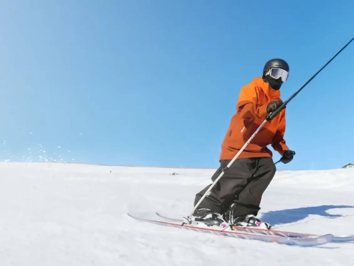 Skieur en veste orange qui descend une piste, perche à selfie tenue à bout de bras