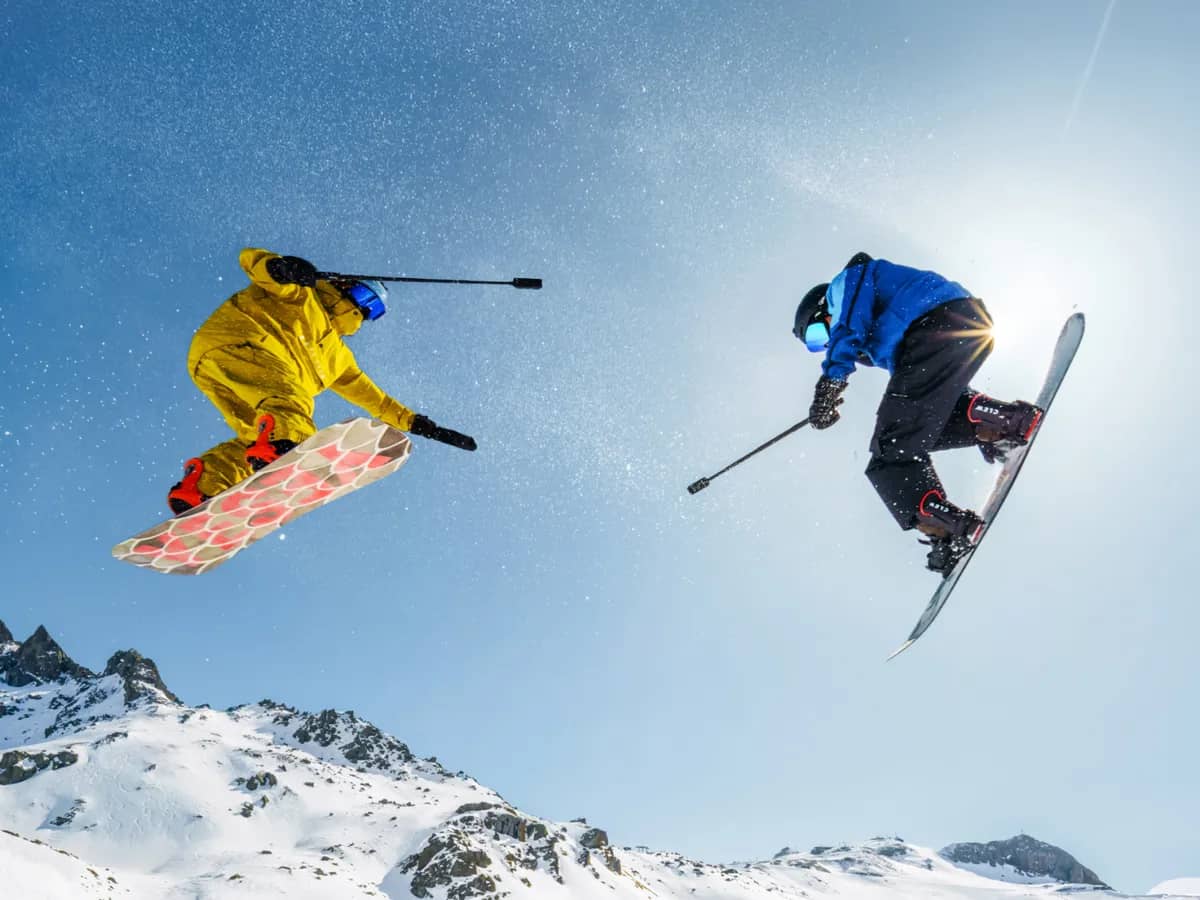 Deux snowboardeurs en plein saut au-dessus de sommets enneigés, perche à la main