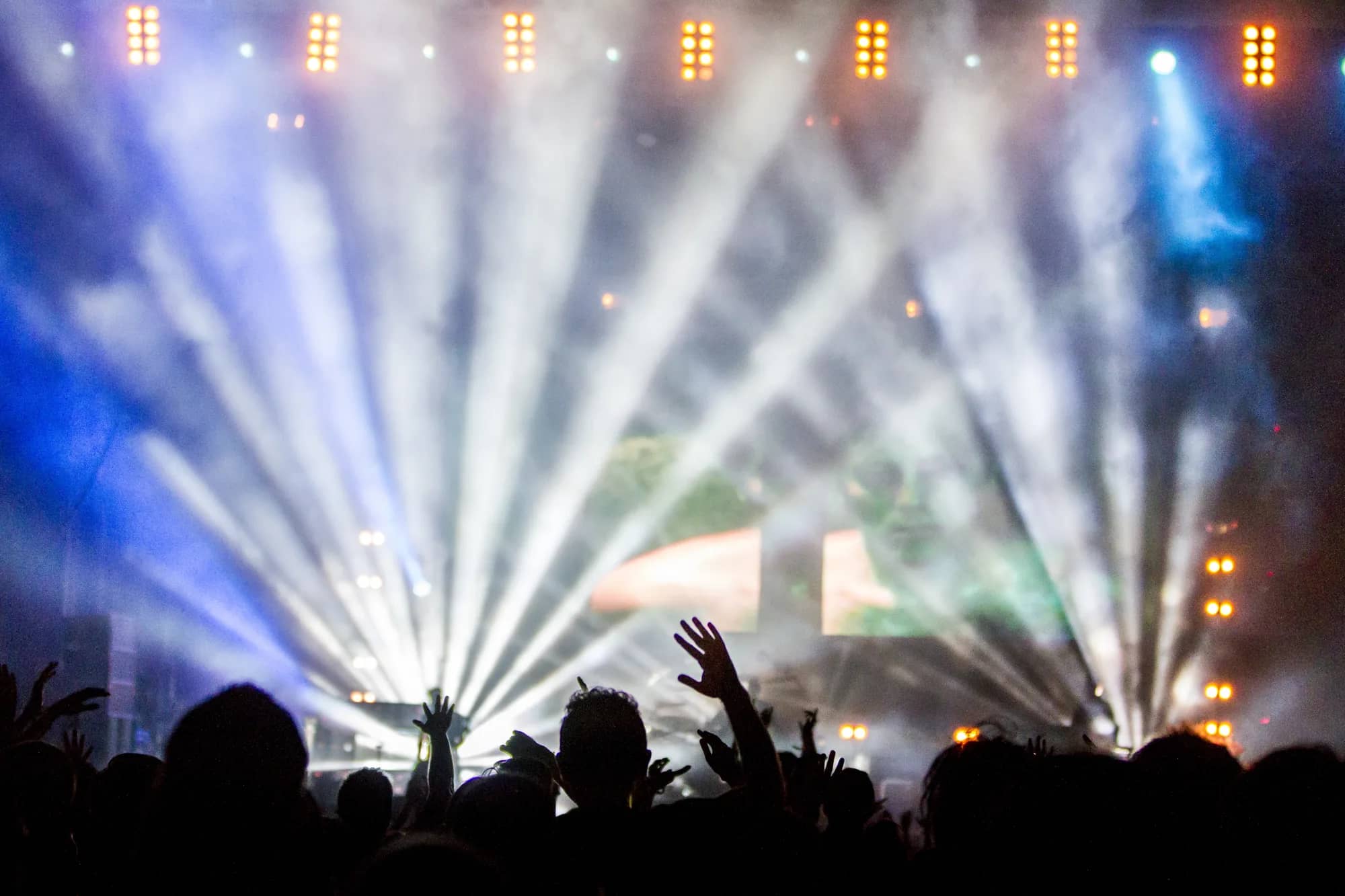 Concert crowd in silhouette, arms raised, under the stage light beams