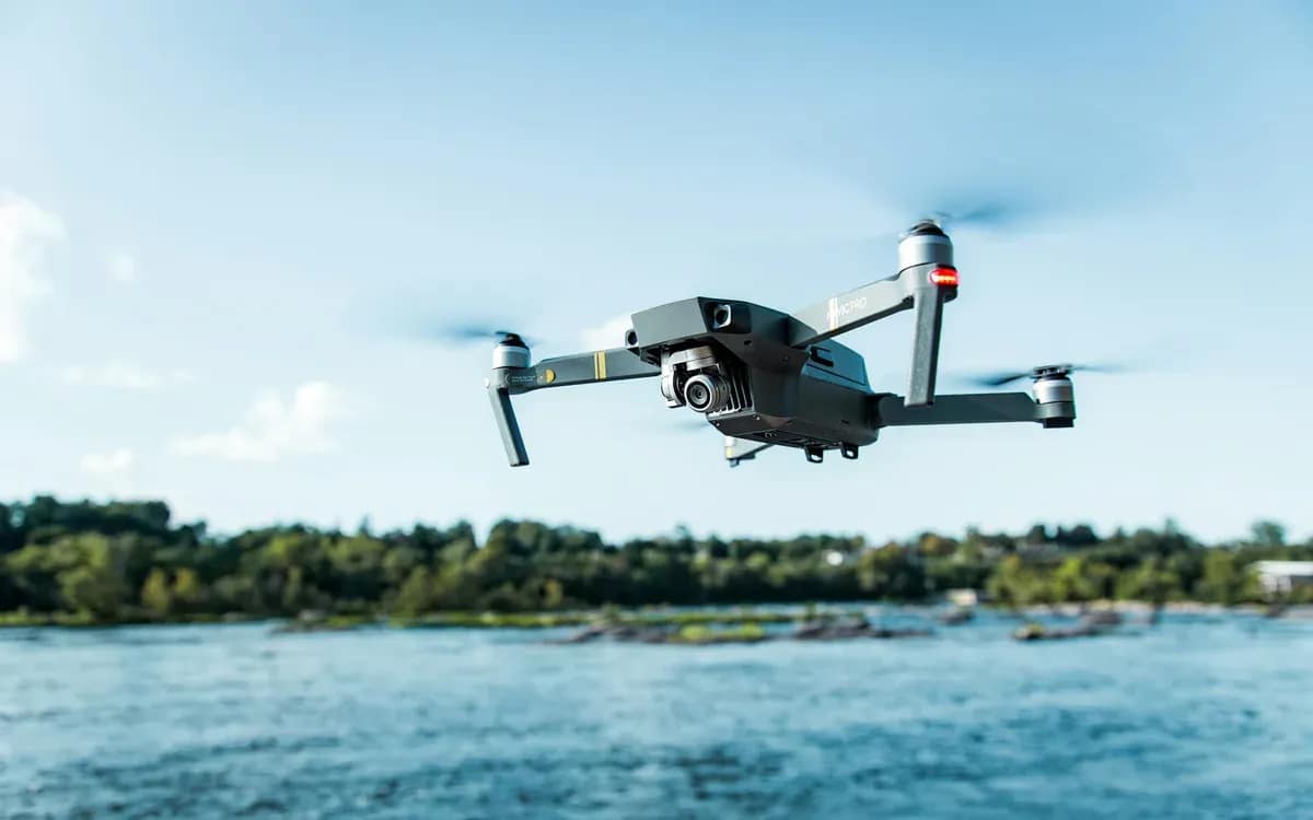 Drone gris en vol au-dessus d’une étendue d’eau bordée d’arbres, sous un ciel bleu