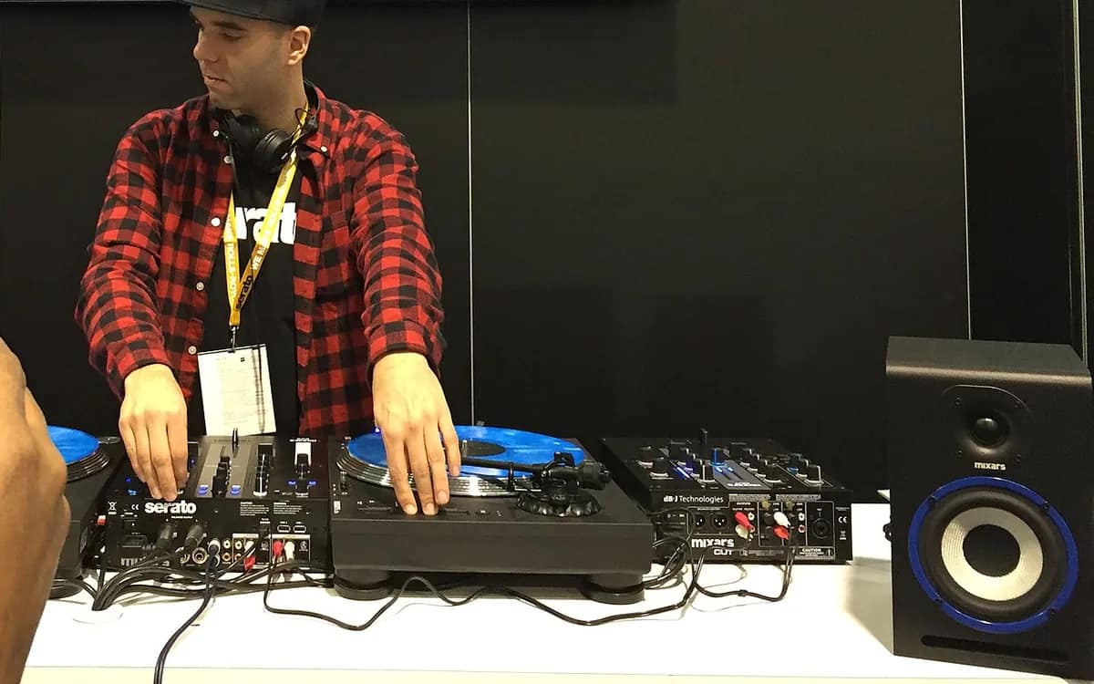 A DJ in a checkered shirt plays on two turntables whose platters bear blue monitor vinyls, on either side of a mixing desk marked Serato.