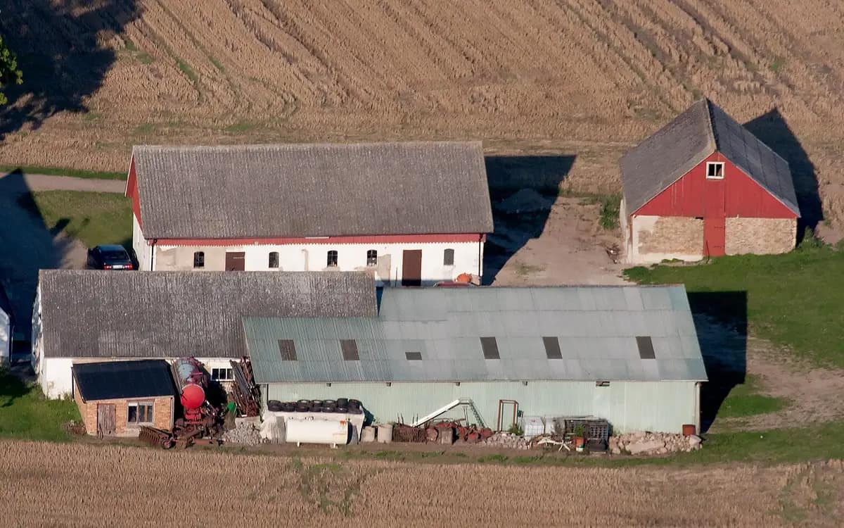 Hoeve vanuit de lucht: een lange schuur met grijs dak, een rode schuur en een plaatstalen loods aan de rand van een geoogst veld