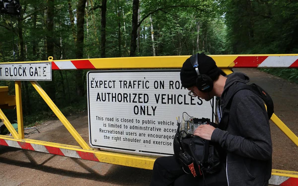 Preneur de son au casque, penché sur son enregistreur de terrain, devant une barrière jaune en lisière de forêt