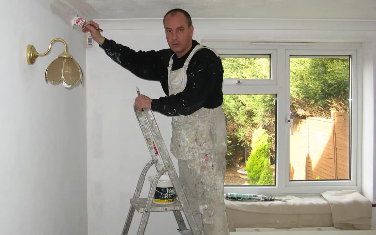 Peintre en combinaison blanche sur un escabeau, rouleau à la main, repeignant l’angle d’un mur près d’une fenêtre