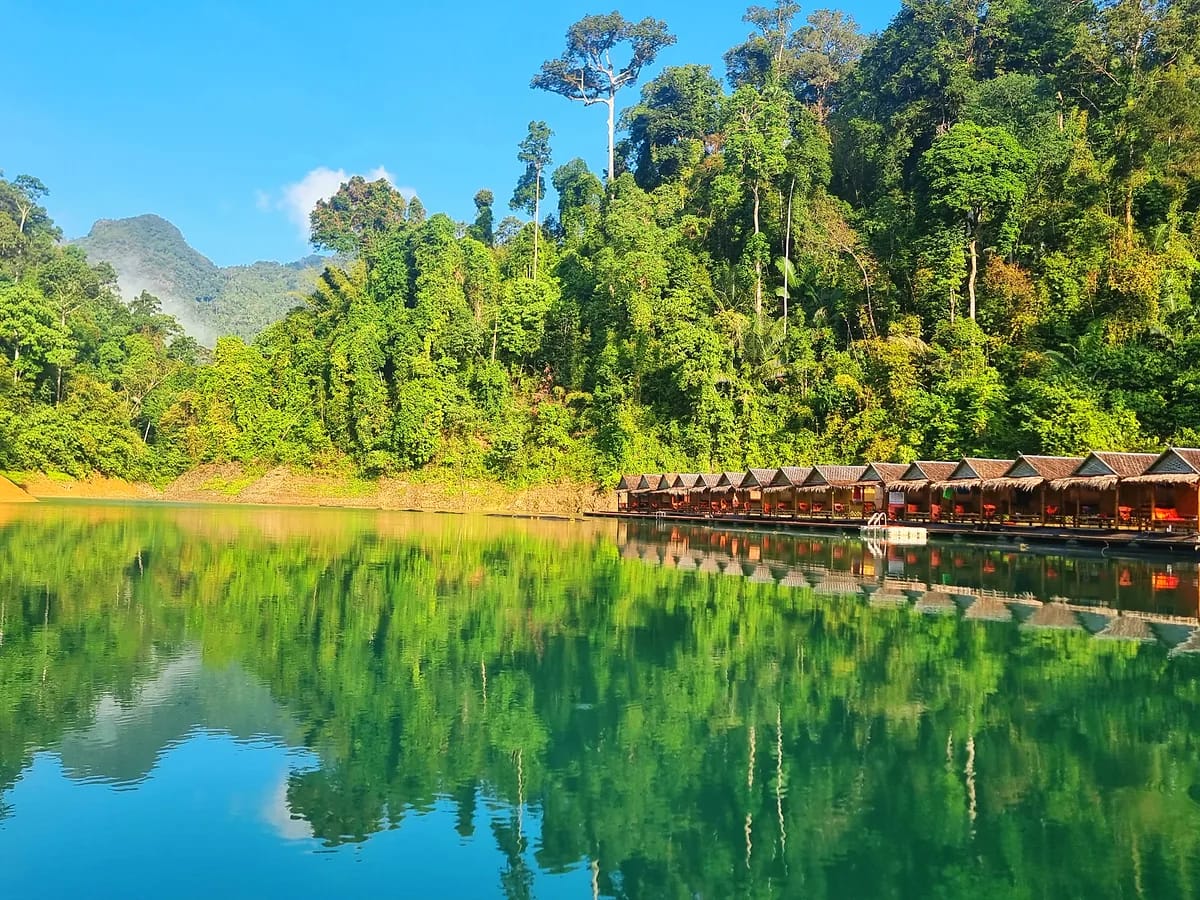 Lac calme bordé de forêt tropicale dense, avec des cabanes flottantes en bois qui se reflètent dans l’eau