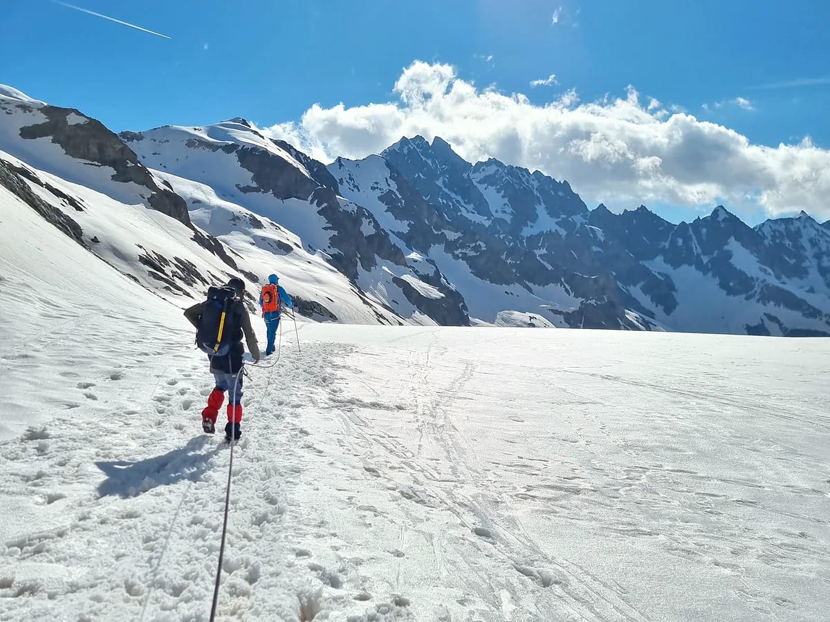 Deux alpinistes encordés progressant de dos sur un glacier enneigé face à des sommets rocheux