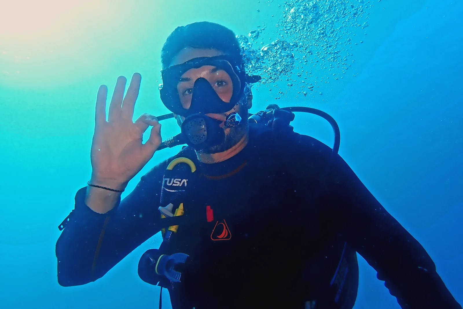 Plongeur en scaphandre faisant le signe OK sous l’eau, dans une mer bleue traversée de lumière