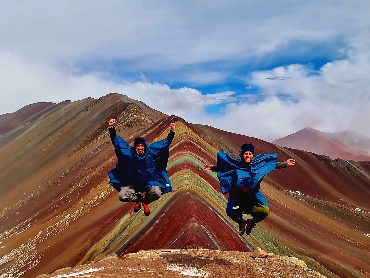 Deux randonneurs en plein saut sur une crête aux roches multicolores, sous un ciel nuageux