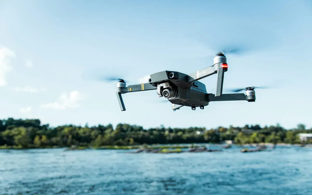 Drone gris en vol au-dessus d’une étendue d’eau bordée d’arbres, sous un ciel bleu