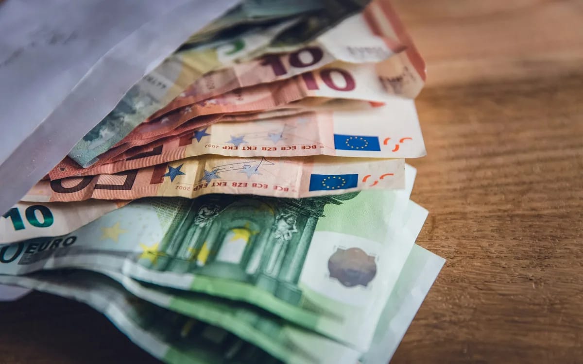 Une liasse de billets de 5, 10, 20 et 50 euros dépassant d’une enveloppe blanche, posée sur une table en bois.