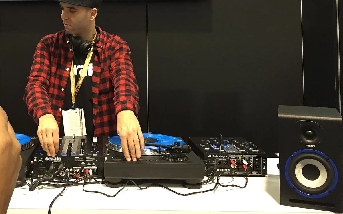 A DJ in a checkered shirt plays on two turntables whose platters bear blue monitor vinyls, on either side of a mixing desk marked Serato.