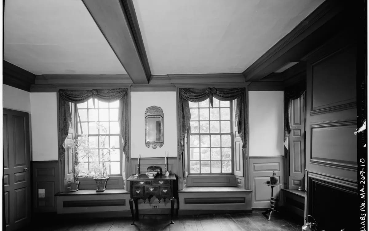 Pièce ancienne photographiée de face, en noir et blanc : deux fenêtres à petits carreaux, lambris et banquettes d’appui