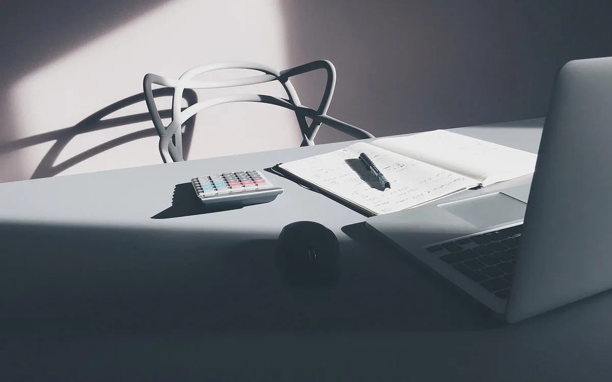 Bureau clair traversé par la lumière : une calculatrice, un carnet ouvert couvert de calculs manuscrits avec un stylo posé dessus, une souris et un ordinateur portable.