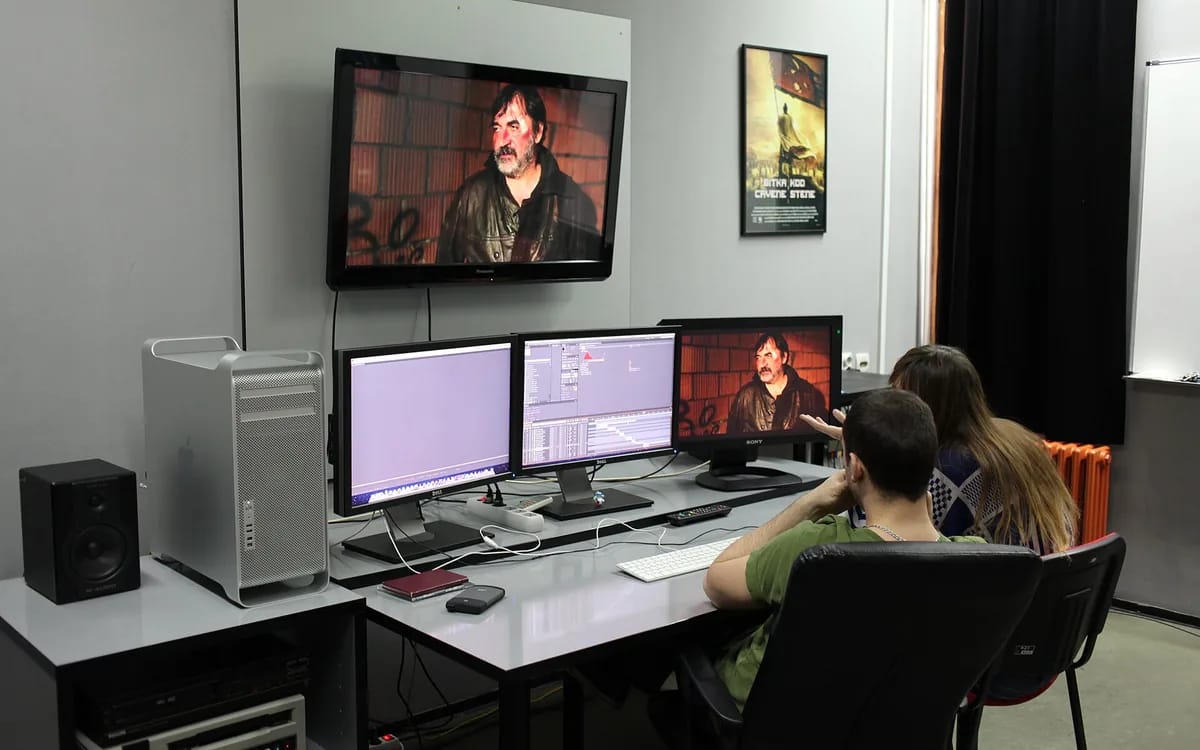 Salle de montage vidéo : une tour d’ordinateur et trois écrans alignés sur un bureau, dont un affiche une timeline de montage, un moniteur de contrôle au mur et deux personnes assises devant.