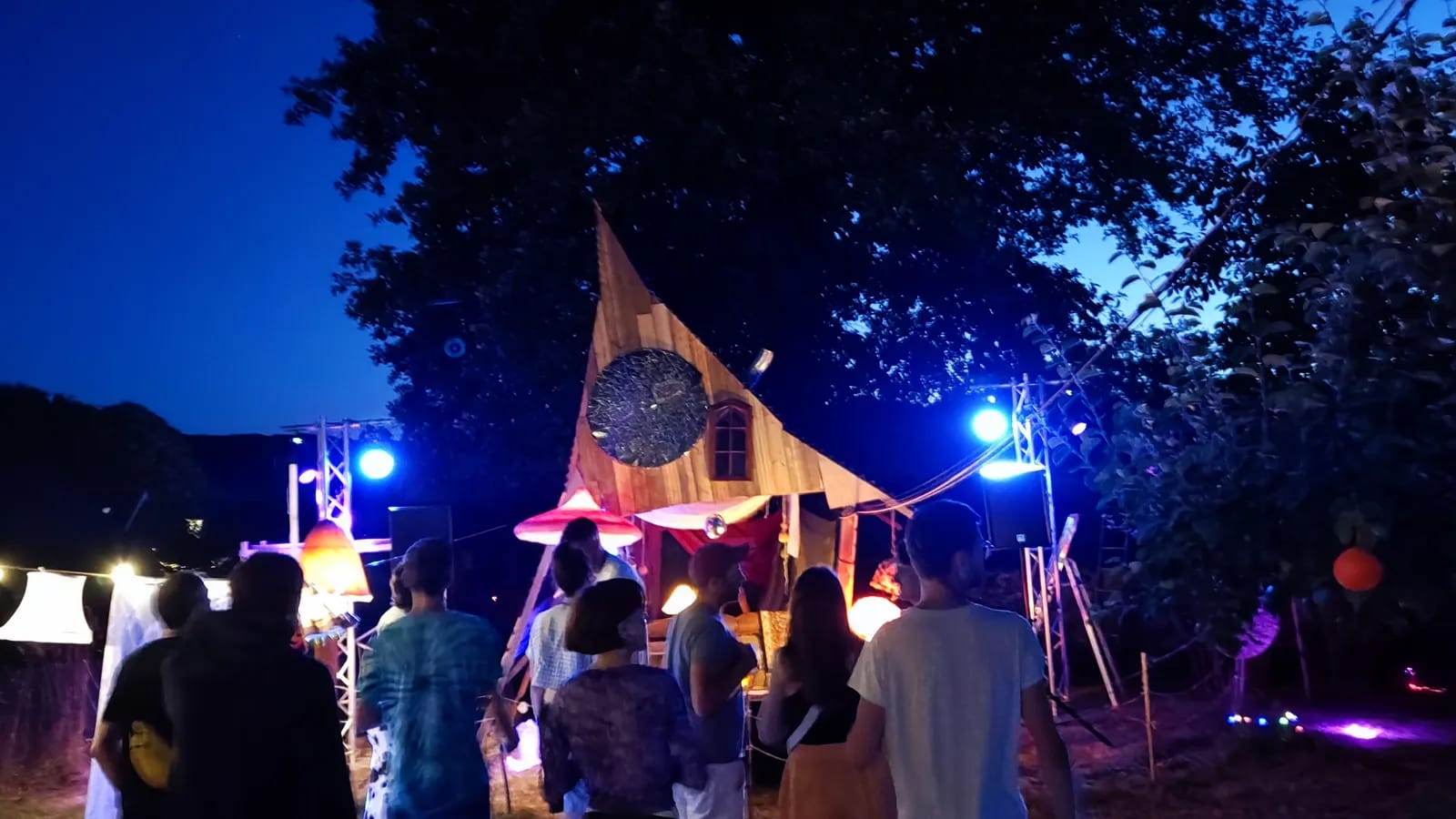 Scène en bois en forme de voile, éclairée en bleu dans un verger à la nuit tombée, devant un public de dos