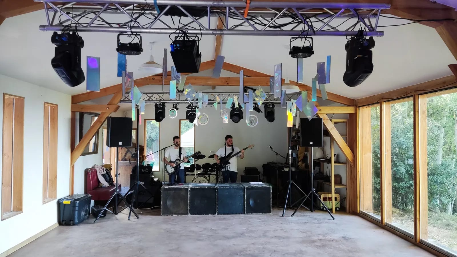Salle claire à charpente de bois : un pont d’éclairage suspendu au-dessus d’une petite scène où jouent deux musiciens