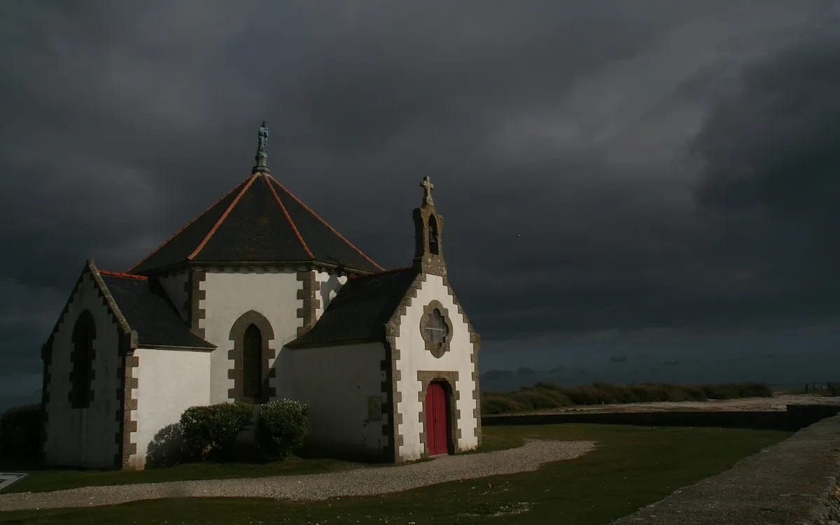 Petite chapelle blanche éclairée par une trouée de soleil sous un ciel d’orage presque noir