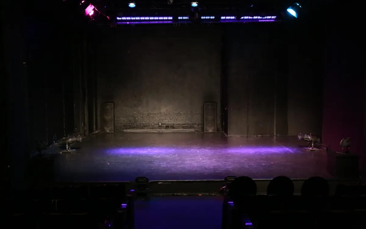 A small empty theatre stage in the dark, lit by a purple glow on the floor