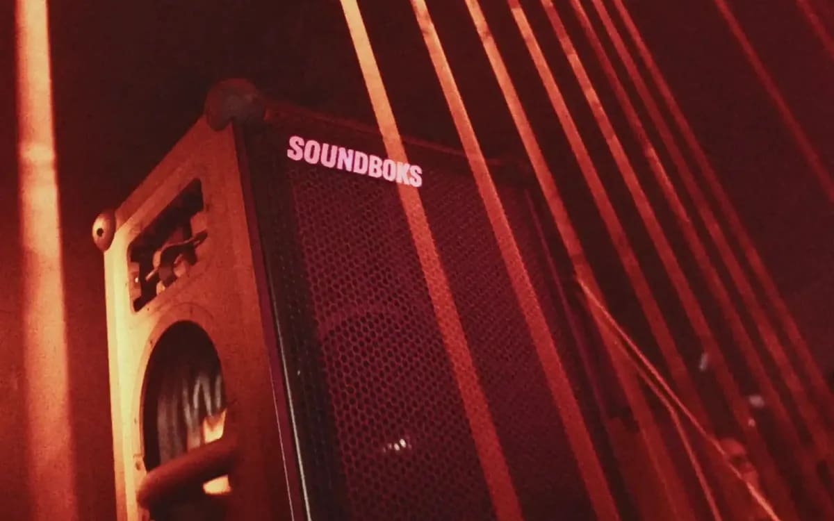 Une enceinte Soundboks 4 posée au sol dans une pièce baignée de lumière rouge, des rayons en arrière-plan