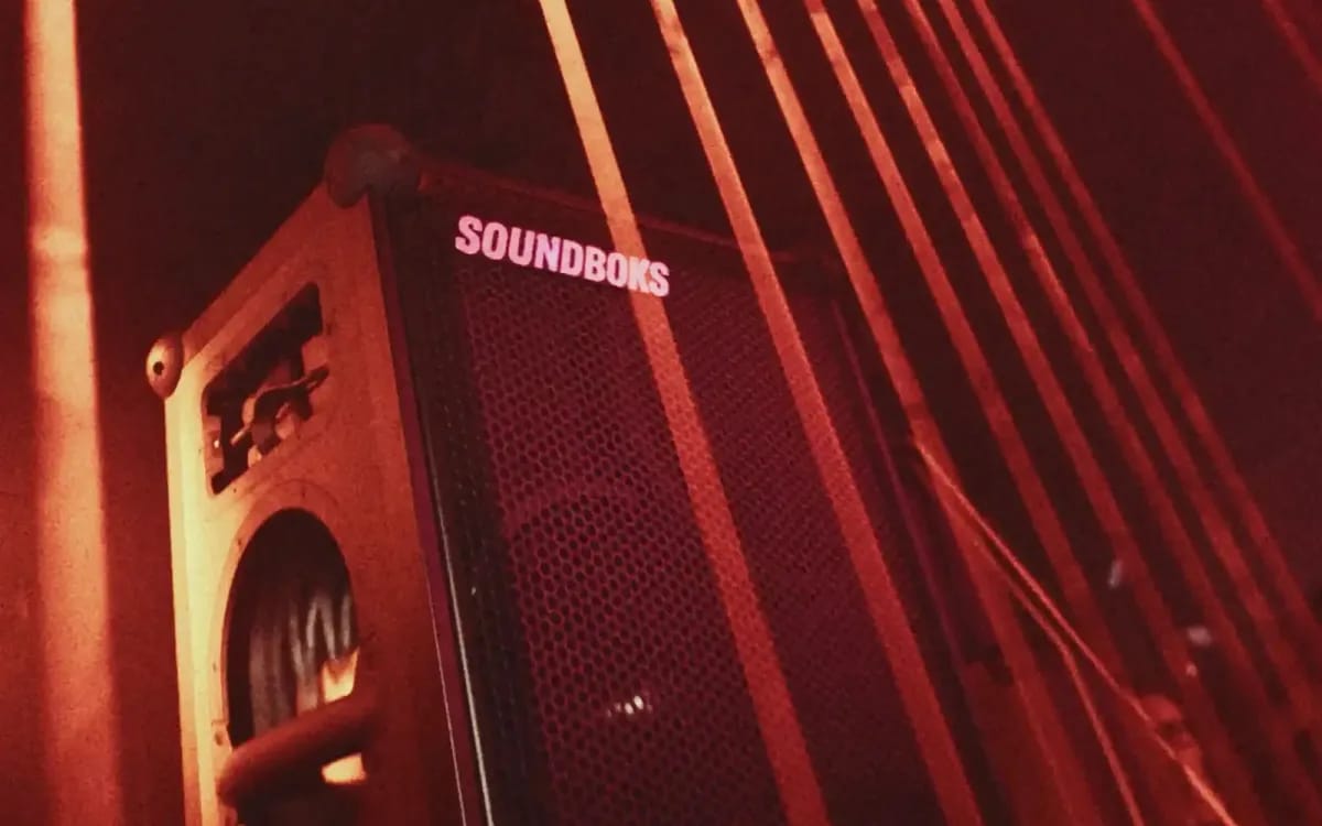Une enceinte Soundboks 4 posée au sol dans une pièce baignée de lumière rouge, des rayons en arrière-plan