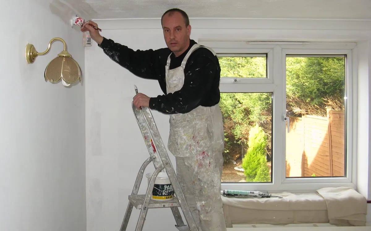Peintre en combinaison blanche sur un escabeau, rouleau à la main, repeignant l’angle d’un mur près d’une fenêtre