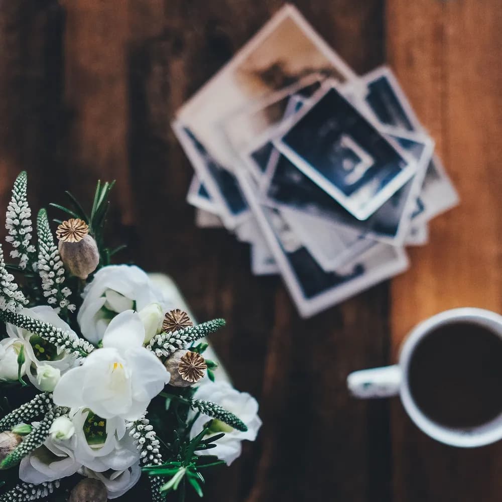Stampe fotografiche appoggiate su un tavolo di legno, accanto a un mazzo di fiori bianchi e a una tazza di caffè