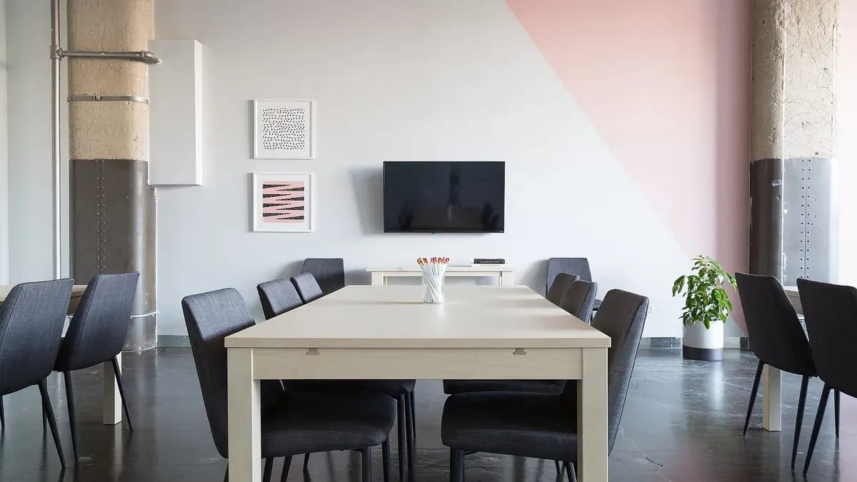 Salle de réunion claire : longue table blanche, chaises noires, écran mural