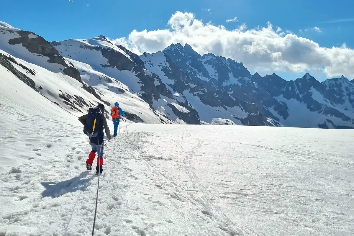 Deux alpinistes encordés progressant de dos sur un glacier enneigé face à des sommets rocheux