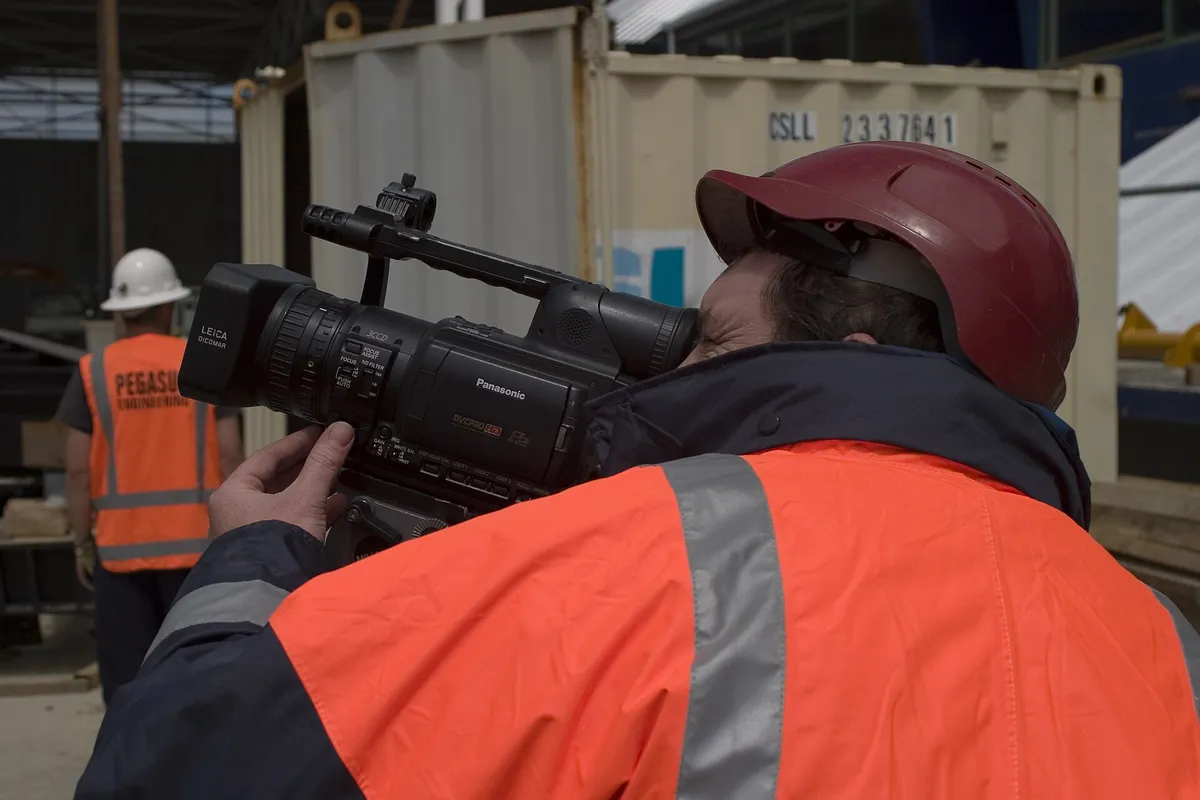 Opérateur en gilet haute visibilité et casque filmant à l’épaule sur un site en activité