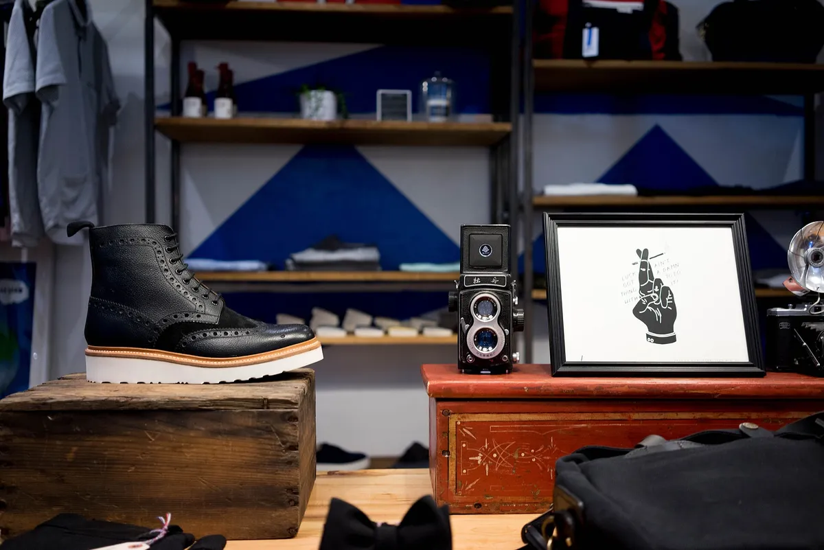 Intérieur de boutique : une bottine et un appareil photo ancien posés en vitrine devant des étagères de vêtements
