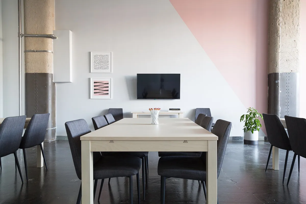 Salle de réunion vide : longue table claire, chaises grises et écran fixé au mur