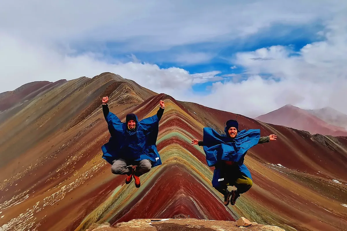 Deux randonneurs en plein saut sur une crête aux roches multicolores, sous un ciel nuageux