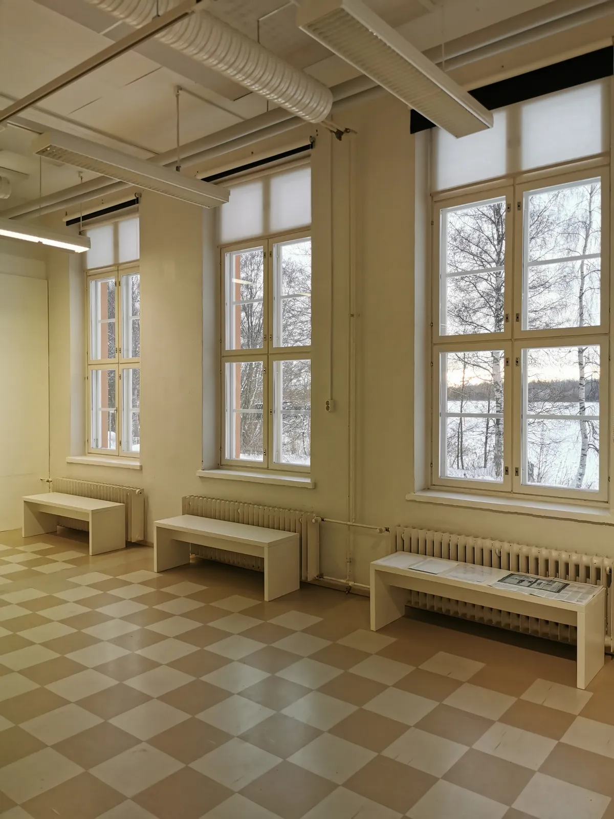 Salle vide aux hautes fenêtres, bancs le long des radiateurs et sol en damier clair, sous une lumière d’hiver uniforme