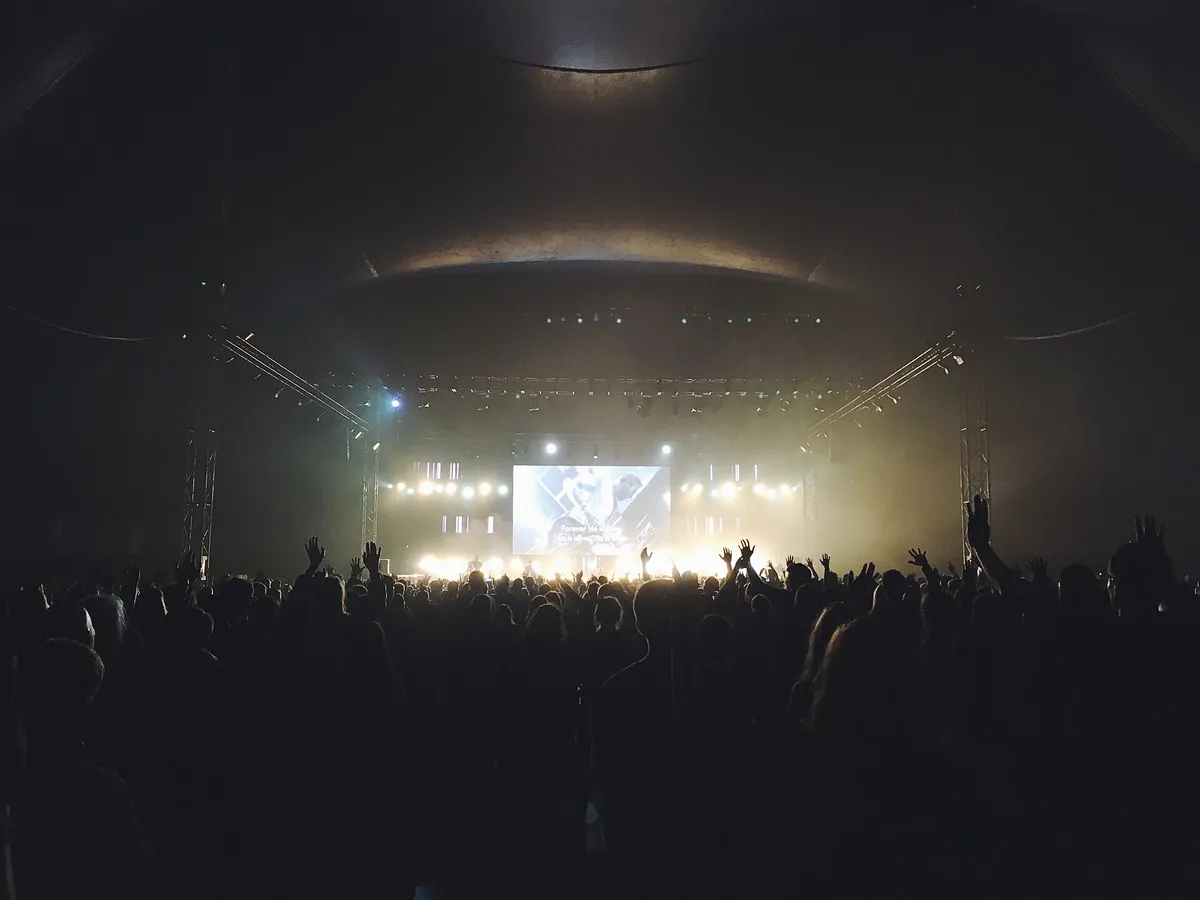 Salle de concert vue du fond : silhouettes du public et scène éclairée au loin
