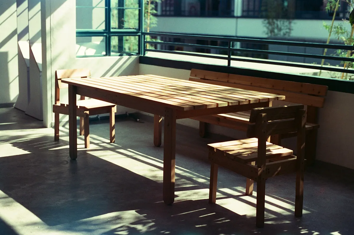 Table et bancs en bois dans une pièce presque nue, sol béton et grande baie vitrée en lumière rasante