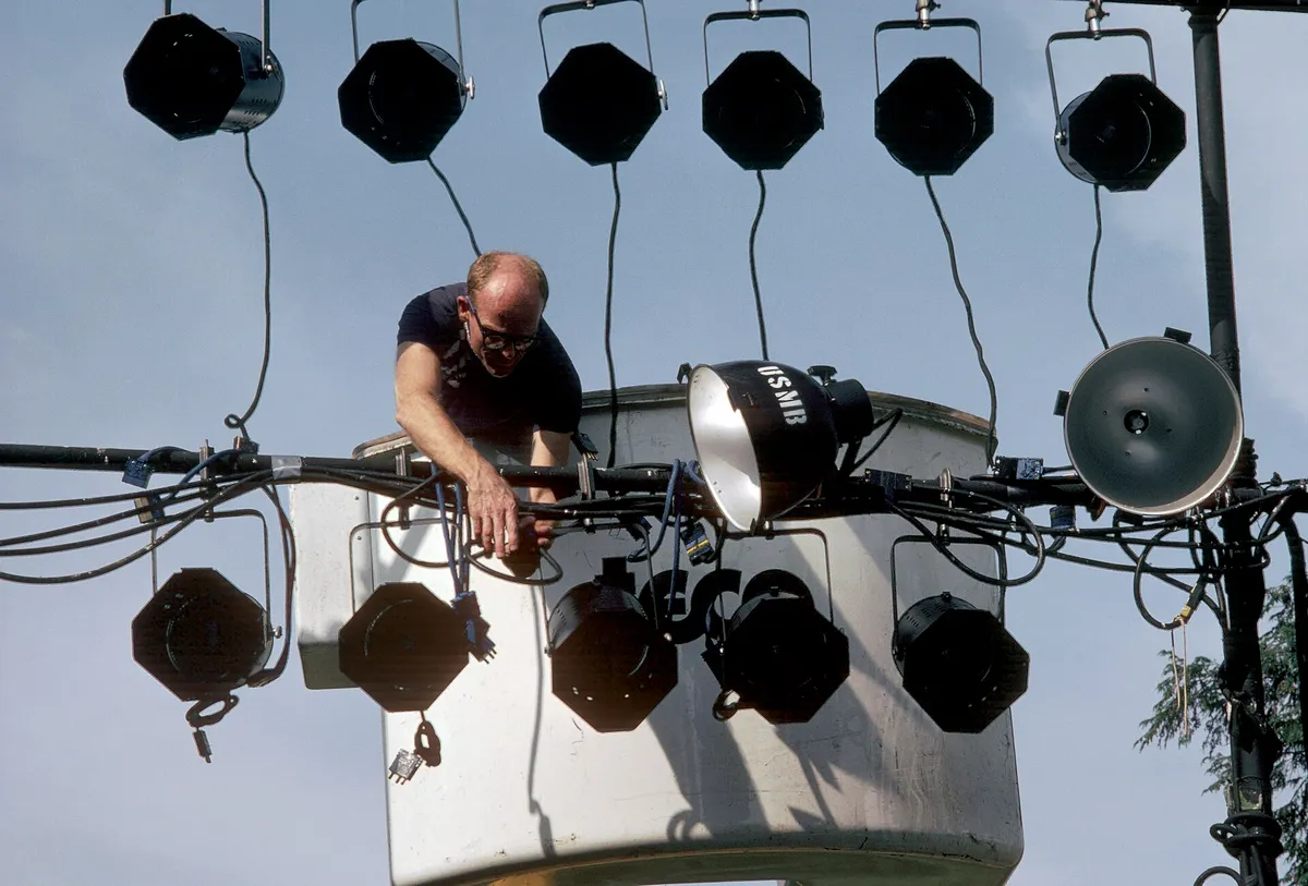 Technicien penché sur une barre de projecteurs de scène, contre un ciel bleu