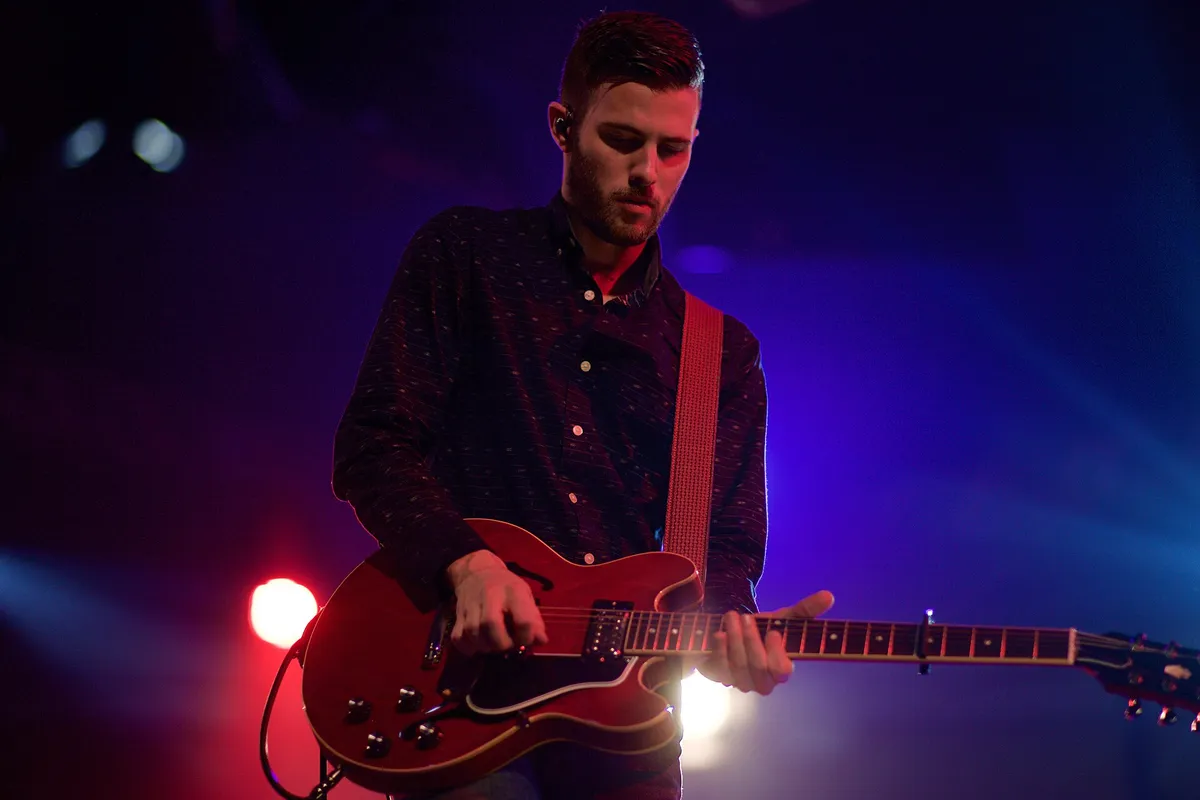 Guitariste sur scène, guitare rouge, fond bleu et violet, éclat rouge au sol