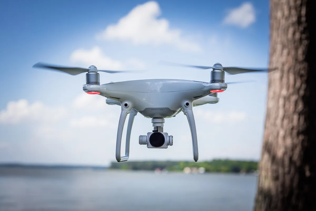 Dron blanc en vol estacionari just al costat d’un tronc d’arbre, cel blau i aigua al darrere