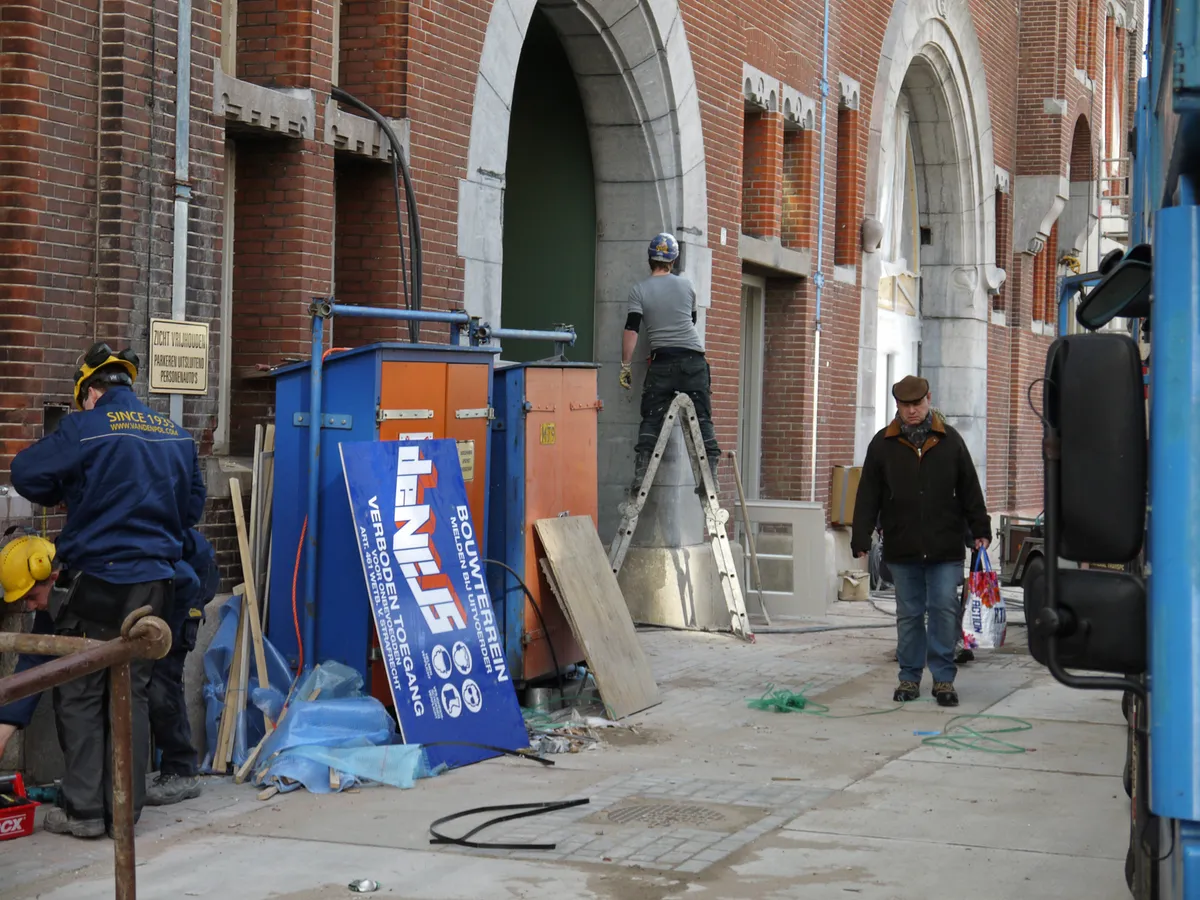 Chantier de rénovation au pied d’un bâtiment en brique : ouvriers, échelle, panneaux de chantier et matériaux au sol