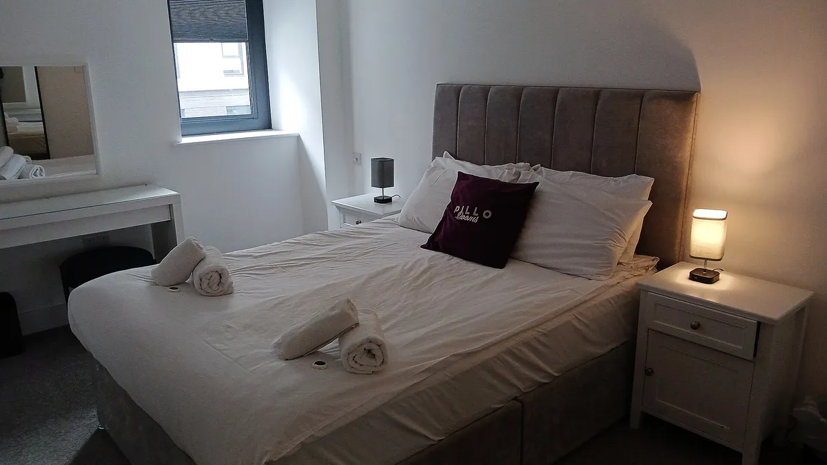 A hotel room with a made double bed, rolled towels and a lit bedside lamp