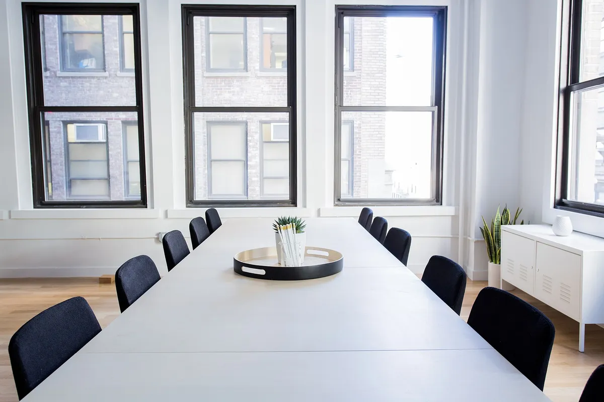 A bright meeting room, a long white table and black chairs before three windows